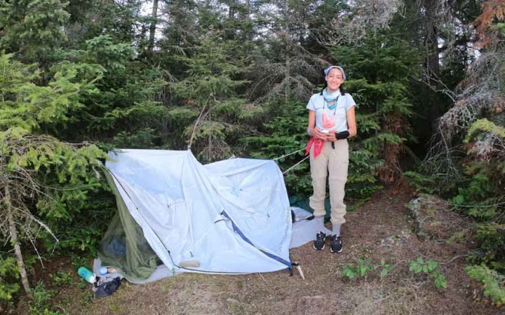an outward bound student stands beside their tarp shelter in a wooded area 